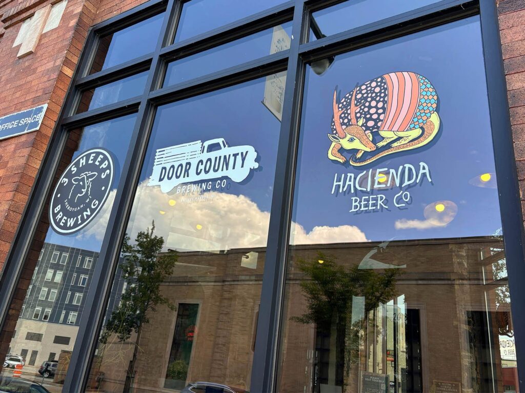 A window with 3 panels. Each panel has a brewery logo. From left to right: 3 Sheeps Brewing, Door County Brewing, and Hacienda Beer Co.